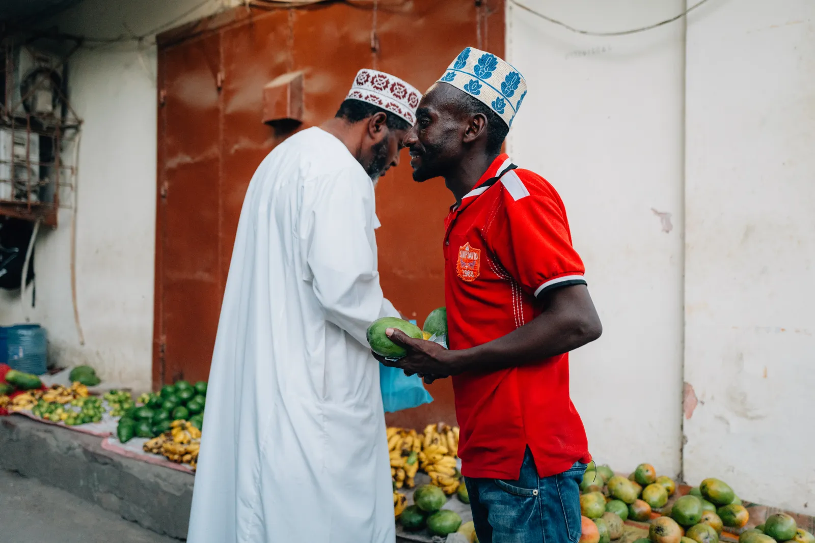 Deux hommes discutant devant un étal de fruits dans une rue, l'un portant un chapeau traditionnel et une chemise rouge.