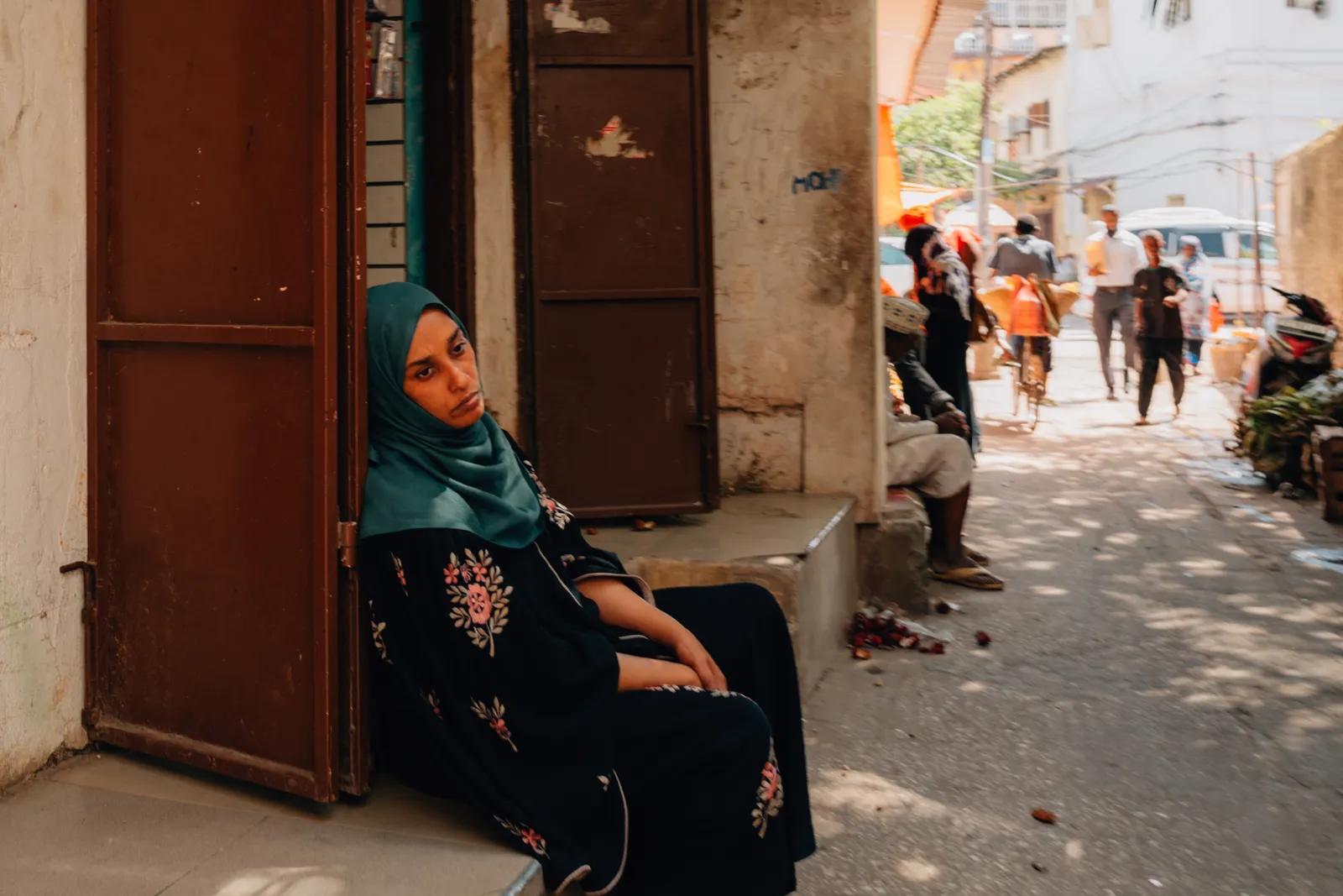 Une femme en tenue traditionnelle assise à l'entrée d'une boutique dans une rue animée de Stone Town.