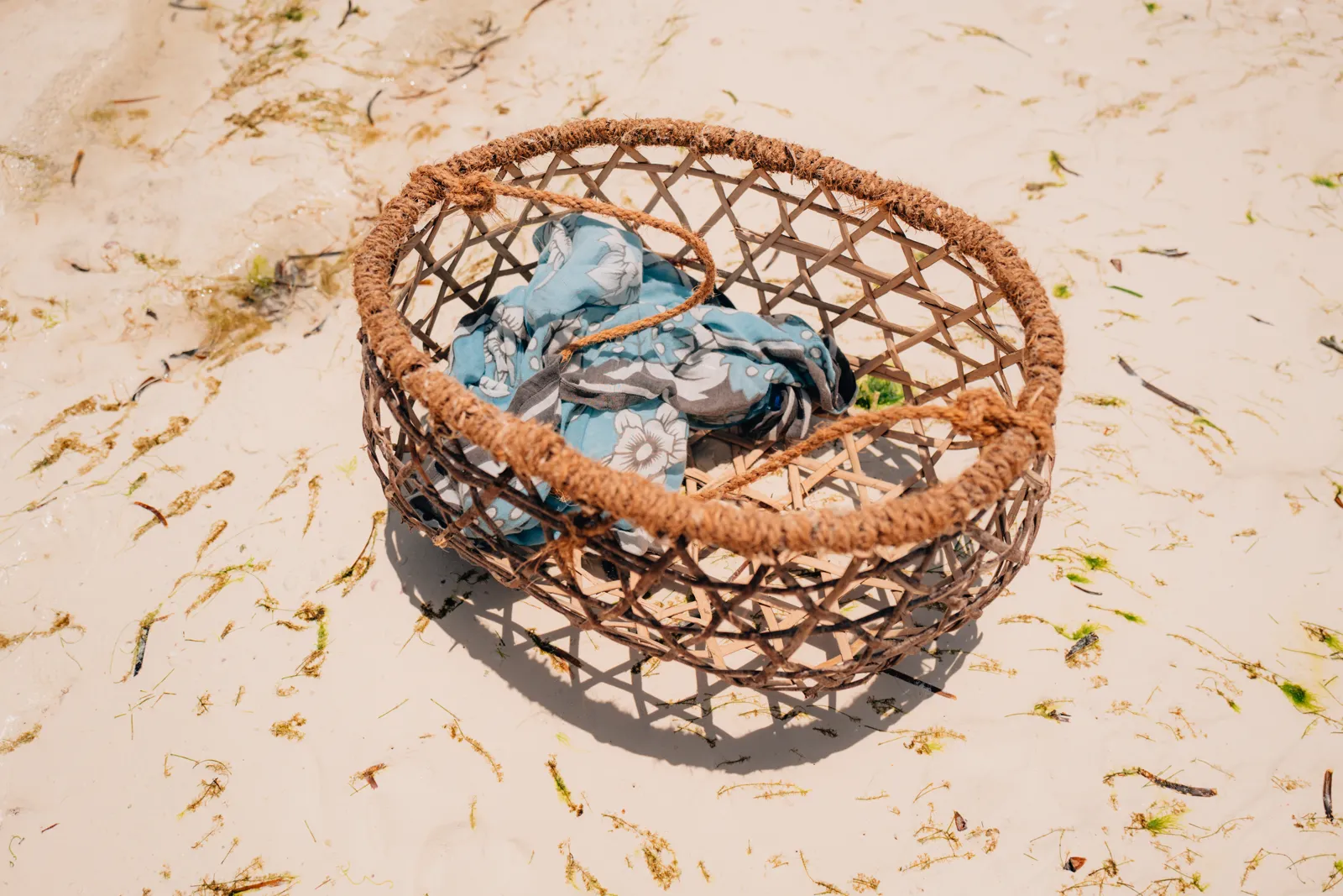 Panier en osier tressé contenant un tissu bleu, posé sur une plage de sable parsemée d'algues.